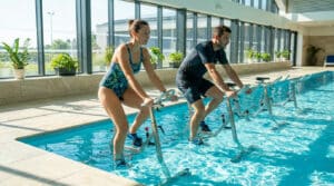 Un homme et une femme font de l'aquabike dans une piscine intérieure moderne et lumineuse. Ils portent des maillots de bain et des chaussures aquatiques.